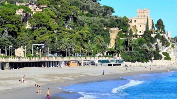 Una playa con un edificio parecido a un castillo al fondo, un muelle que se adentra en el agua, y gente disfrutando de la playa.