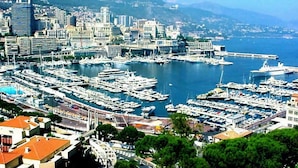 Un puerto deportivo con numerosas embarcaciones amarradas, rodeado por un paisaje urbano con edificios modernos y montañas al fondo.