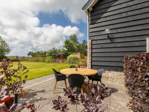 An outdoor patio with a round wooden table and four chairs, a brick wall, and a garden with a wooden fence in the background.