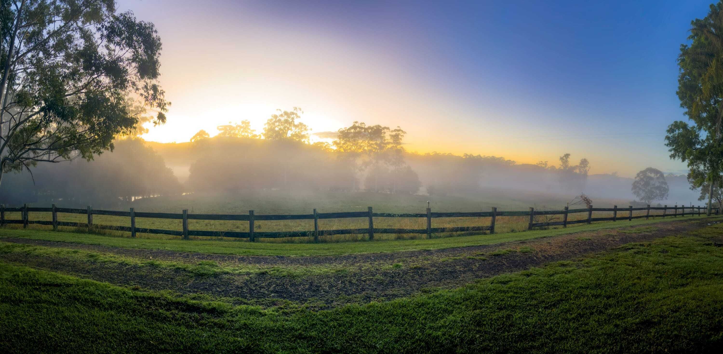 Paisaje rural con valla de madera, árboles y un amanecer o atardecer.