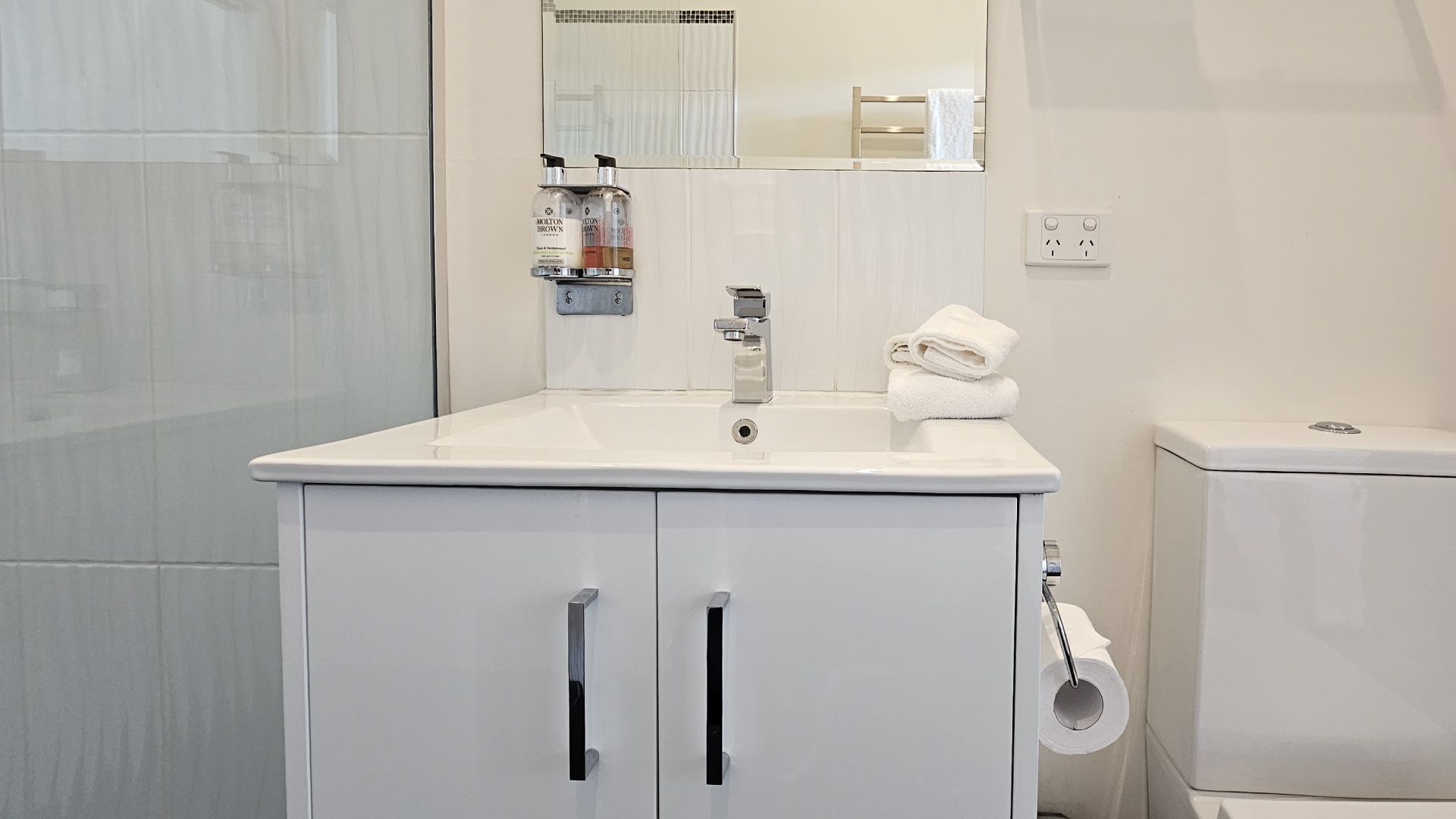 Ein Badezimmer mit einem weißen Waschbecken, Spiegel und WC. Unter dem Waschbecken befinden sich zwei Schränke, einer davon mit einem Handtuchhalter.