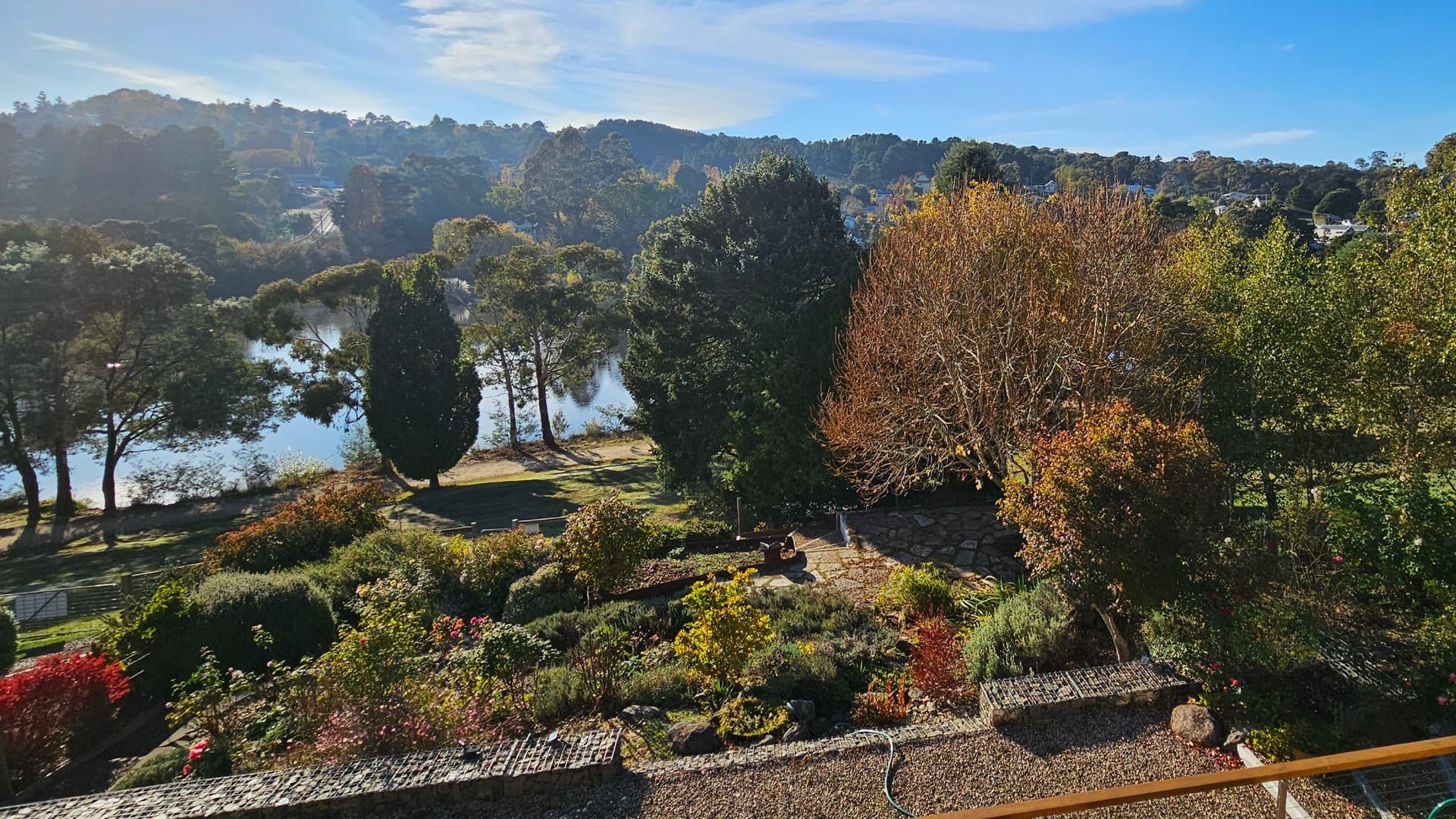 Ein angelegter Garten mit verschiedenen Bäumen und Sträuchern, einem Kiesweg und einem Blick auf einen See und Hügel im Hintergrund.