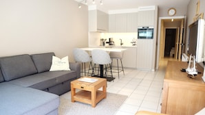 Ein modernes Wohnzimmer mit einem grauen Sofa, zwei Sesseln, einem Holztisch und einer Küchenzeile mit Esstisch und Stühlen.