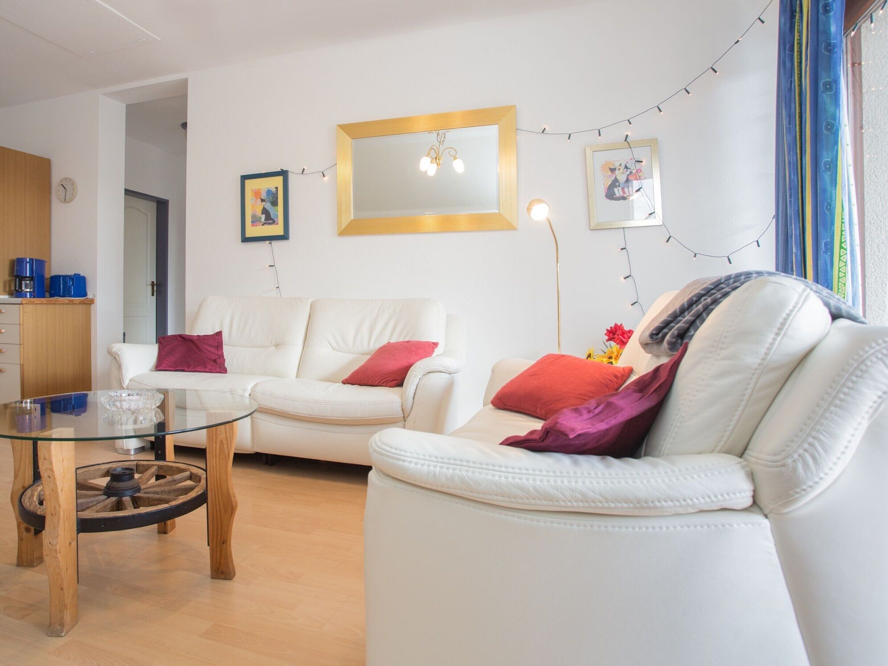 Una sala de estar moderna con un sofá blanco, una mesa de centro de vidrio y una mesita de noche de madera. Hay dos cuadros en la pared, un reloj y una guirnalda de luces.