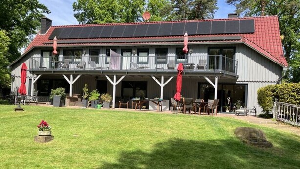 A two-story building with a red roof, a balcony with red umbrellas, and a grassy lawn with potted plants. - Lovely Apartment in Neustadt (Neustadt am Ruebenberge)