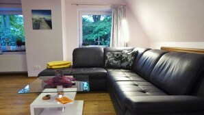 A living room with a black leather sofa, a glass coffee table, and a view of greenery through a window. - Lovely Apartment in Neustadt (Neustadt am Ruebenberge)