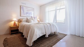 A bedroom with a bed, nightstands, a wall-mounted power outlet, and a large window with sheer curtains. - Cosy, fully-renovated apartment with ocean views and 1-minute walk to the beach (West Beach)
