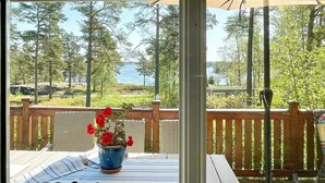 A covered outdoor area with a wooden deck, a wicker table with a striped cloth, and a potted plant with red flowers. - 5 Person Holiday Home in Gunnebo (Gunnebo)