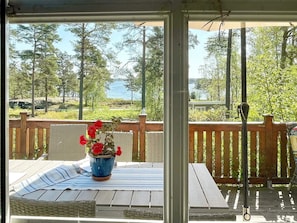 Un espace extérieur abrité, doté d’une terrasse en bois, d’une table en osier recouverte d’une nappe à rayures et d’un pot de fleurs aux fleurs rouges.