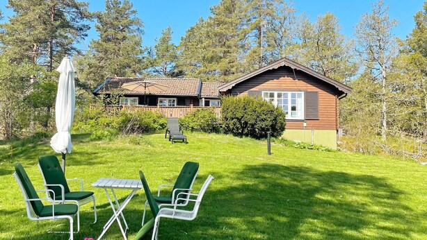 A house with a wooden exterior, a lawn with green chairs and a table, and a white umbrella. - 5 Person Holiday Home in Gunnebo (Gunnebo)