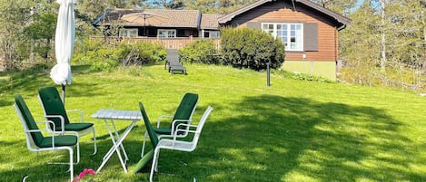 Una casa con fachada de madera, un césped con sillas verdes y una mesa, además de un paraguas blanco.