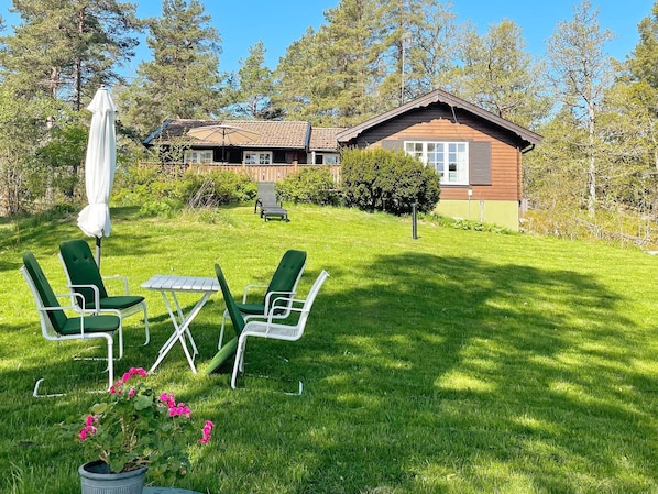 Une maison avec une façade en bois, une pelouse avec des chaises vertes et une table, et un parasol blanc.