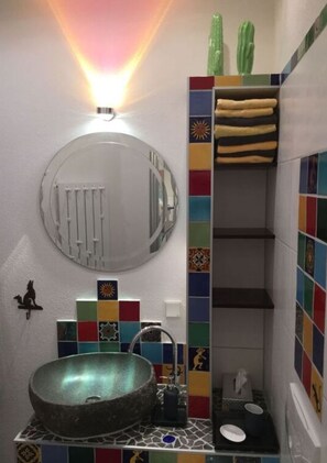 A bathroom with a round mirror, a stone sink, colorful tiles, a cactus decoration, and a shelf with rolled towels. - Holiday Apartment Anderewelt-bochum (Bochum)