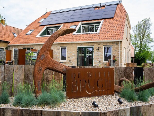 A building with a sign reading "De With Bruch Appartements" surrounded by a wooden fence and greenery.