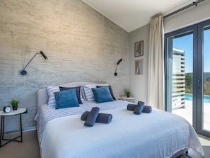 Un dormitorio moderno con una cama grande, dos mesitas de noche, una pequeña planta en maceta y vistas a una piscina a través de un ventanal corredero.