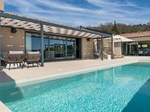 Un área de piscina moderna al aire libre con un porche cubierto, asientos y una vista despejada del paisaje circundante.