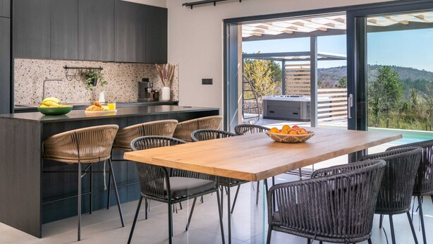 Una cocina moderna con armarios oscuros, una isla central y un comedor con sillas de mimbre.