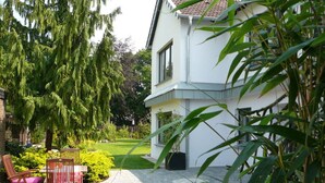 Ein zweistöckiges Haus mit gepflasterter Terrasse, einem Holzstuhl und einem großen Baum.