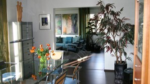 Ein Essbereich mit Glastisch, Stühlen und einem Blumenstrauß. Im Hintergrund befindet sich eine Couch und ein Gemälde.