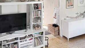 A modern living room with a large flat-screen TV, a white bookshelf filled with books and decorative items, a wooden coffee table, and a white cabinet with a sink. - 5 Person Holiday Home in Djurhamn-by Traum (Djurhamn)