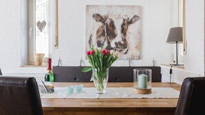 En matplats med ett träbord, två stolar, en vas med blommor, en flaska och en tavla med kor på väggen.