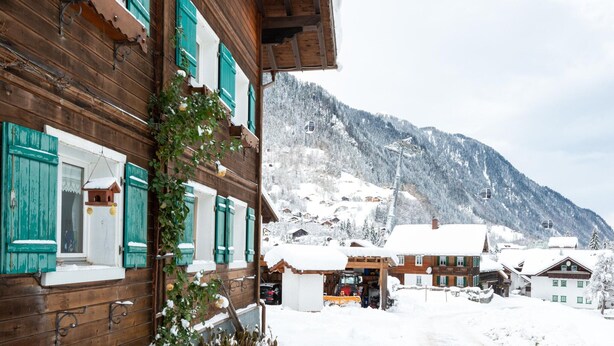 Un bâtiment en bois avec des volets verts, une montagne enneigée en arrière-plan et un télésiège visible.