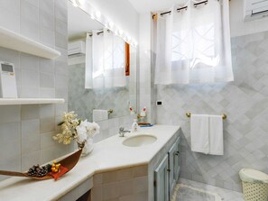 Une salle de bain moderne avec un lavabo blanc, un meuble-lavabo et un grand miroir avec éclairage intégré. Deux serviettes blanches sont accrochées au mur, ainsi qu’une petite poubelle et une plante décorative posée sur le comptoir.