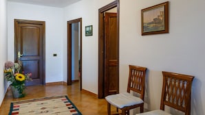 Um corredor com porta de madeira, um quadro pendurado na parede e duas cadeiras de madeira com almofadas.