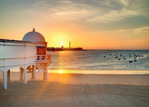 Um edifício costeiro com cúpula e varanda, com vista para uma praia com barcos e um farol ao longe.
