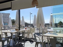 Un espace de restauration en plein air avec des tables et des chaises, des parasols et une vue sur un bâtiment moderne et des palmiers.