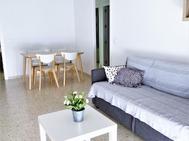 A modern living room with a grey sofa, a white coffee table with a plant, and a dining area with wooden chairs and a table.