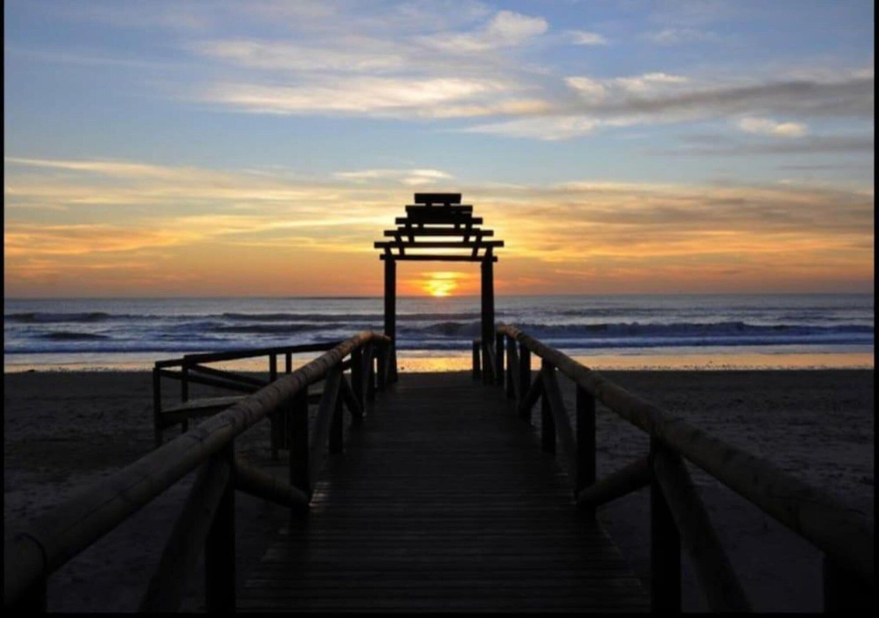A wooden pier leading to a structure with a tiered roof, overlooking the ocean at sunset.