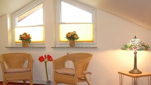 Une pièce avec deux fauteuils en osier, une petite table avec une lampe et deux fenêtres ornées de jardinières.