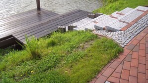 Une jetée en bois s’étendant dans une rivière paisible, avec une zone herbeuse et un chemin de pierre menant à un bateau amarré.