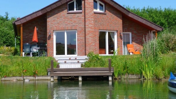Une maison en briques avec un toit en pente, de grandes fenêtres et une jetée qui s’avance dans un plan d’eau.