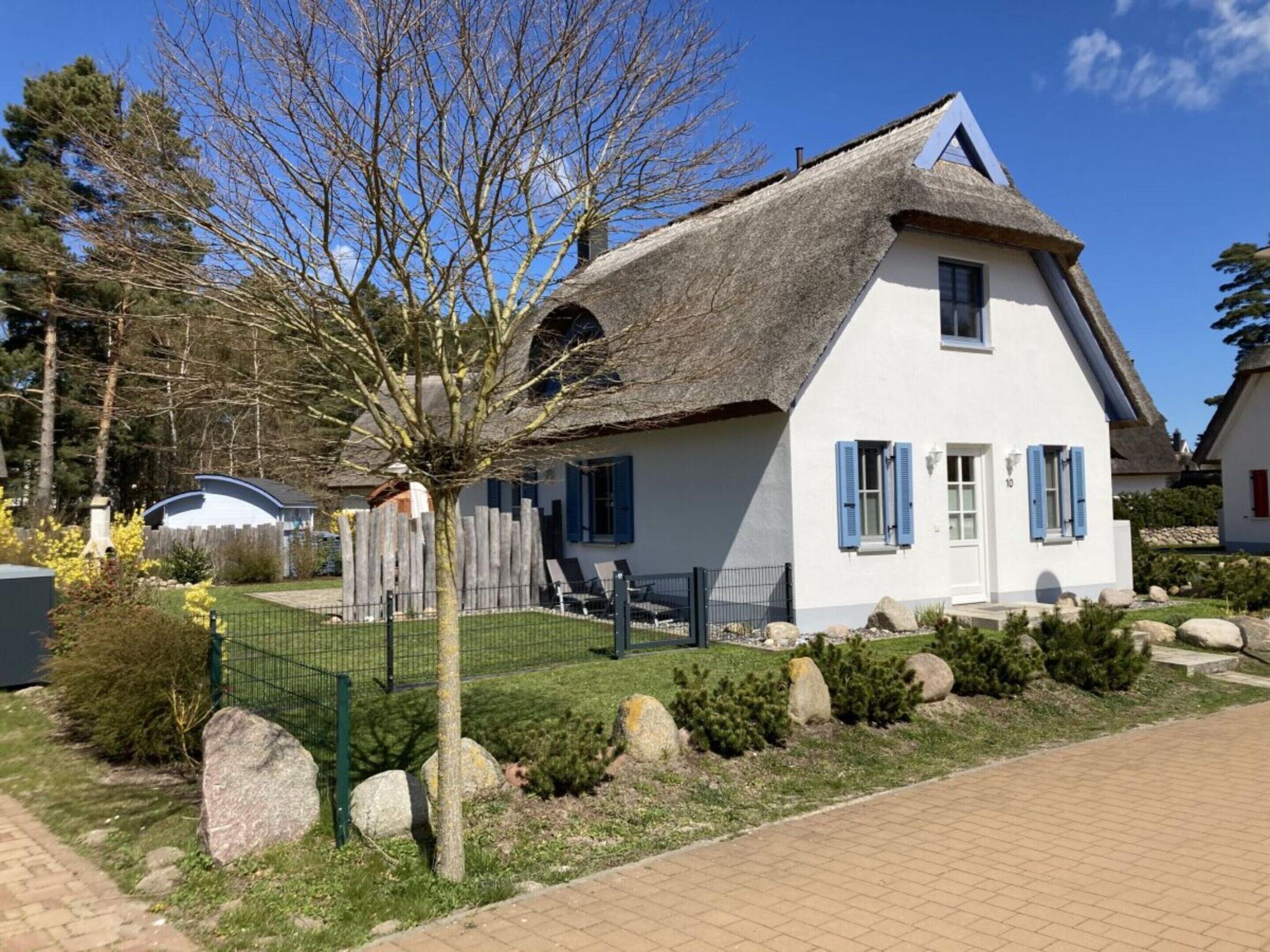 Uma casa branca com telhado de palha, persianas azuis e um jardim cercado por pedras e arbustos.
