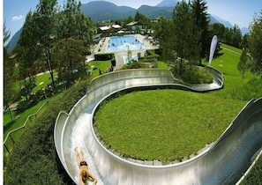 Un tobogán de agua con piscina al final, rodeado de follaje y montañas al fondo.