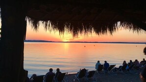 A beach scene with people relaxing on lounge chairs under a thatched roof, overlooking a calm sea at sunset. - Holiday Apartment Dosbach (Reit im Winkl)