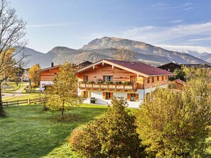 Un chalet en bois à deux étages, doté d’un balcon, entouré d’une végétation luxuriante et avec des montagnes en toile de fond.