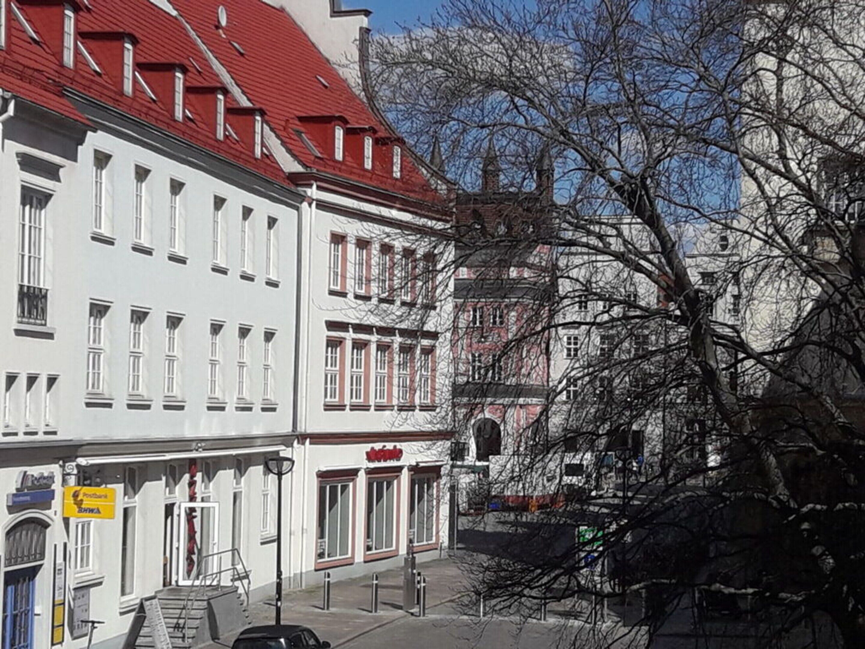 Vista de la calle con un edificio blanco con detalles rojos y un techo rojo, un letrero de 'Postbank', y un árbol con ramas desnudas.