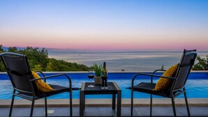 Une vue sur la piscine, avec deux chaises et une table, donnant sur une étendue d’eau et un paysage lointain au coucher du soleil.