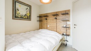 A bedroom with a bed, a wall-mounted shelf with books, a framed painting, and a hanging light fixture. - Beautiful Apartment on the sea Wall (Blankenberge)