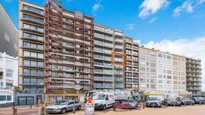 A multi-story building with a construction site on the left, a parking area with cars, and a clear blue sky. - Beautiful Apartment on the sea Wall (Blankenberge)