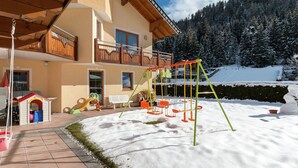 Un edificio a più piani con un balcone in legno, un'area giochi per bambini con altalene e scivoli, e un paesaggio innevato con alberi.