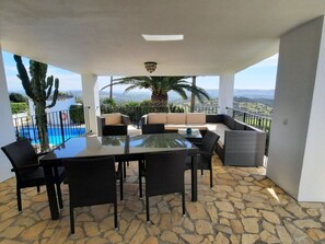 A balcony with a glass table and chairs, a sofa, and a view of a pool and landscape.