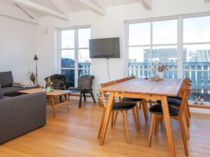 Una sala de estar moderna con un sofá, una mesa de comedor con sillas, un televisor montado en la pared y ventanas amplias.