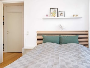 Un dormitorio con cama, cabecera de madera, dos almohadas, pared blanca, estante con cuadros enmarcados y una puerta.