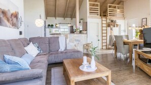Un vaste espace de vie doté d’un plafond en bois, d’un canapé gris, d’une table basse en bois et d’une cuisine à l’arrière-plan.