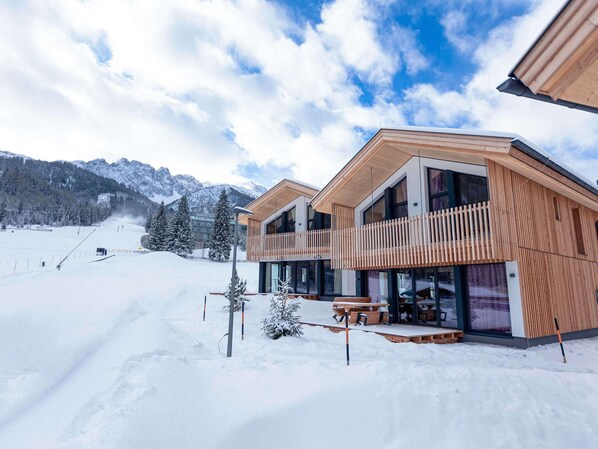 A modern chalet with a wooden exterior, large windows, and a balcony overlooking a snowy landscape with mountains in the background.