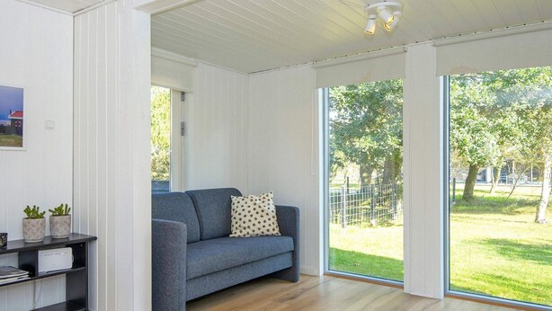A modern living room with a grey sofa, a wooden floor, and large windows offering a view of a grassy area and trees. - 4 Person Holiday Home in Ulfborg (Ulfborg)